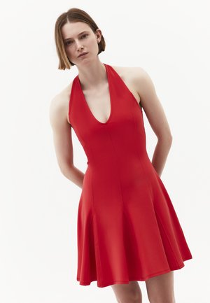 Donna con capelli castani corti che indossa un vestito rosso senza maniche con collo all'americana, in piedi davanti a uno sfondo bianco semplice.