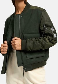 Donkergroene bomberjack met een wollen lichaam, glanzende nylon mouwen, geribbelde manchetten, rits aan de voorkant en een utility pocket met rits op de linkermouw.