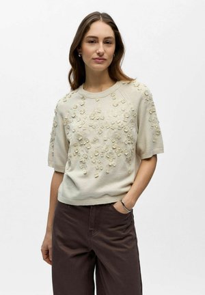 Frau mit langem braunem Haar, die einen cremefarbenen Kurzarm-Pullover mit erhabenen floralen Details und dunkelbraune Hosen trägt, steht mit einer Hand in der Tasche.