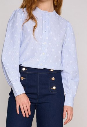 Vrouw draagt een lichtblauw gestreepte blouse met witte geborduurde stippen en donkere, hooggetailleerde broek met gouden knopen.