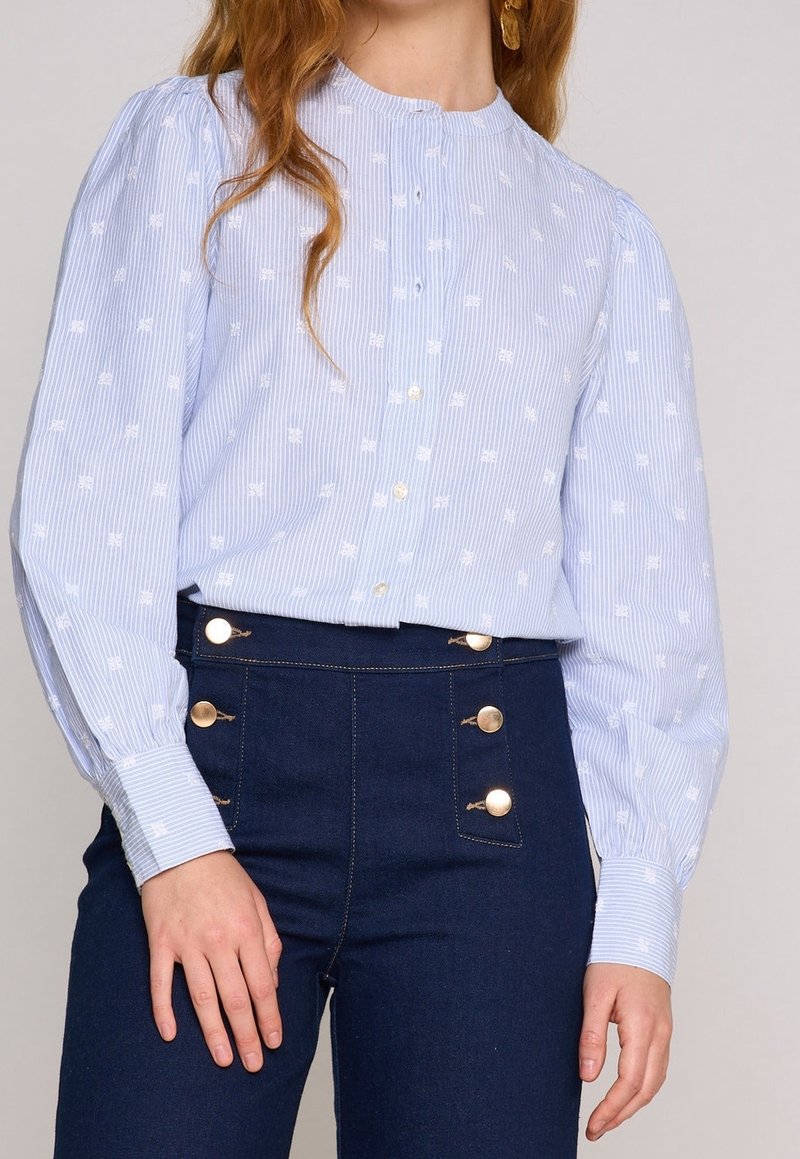 Femme portant un chemisier rayé bleu clair avec des points brodés blancs et un pantalon taille haute foncé avec des boutons dorés.