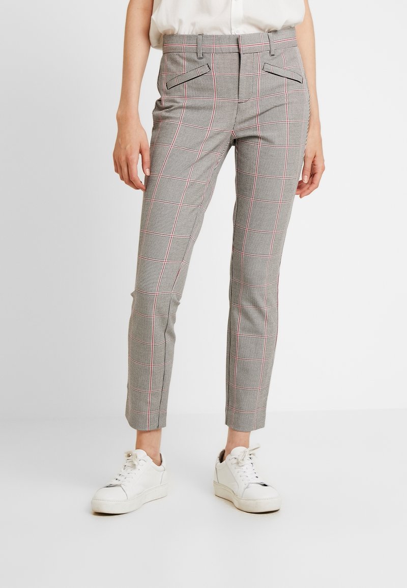 Pantalon gris ajusté à carreaux avec des lignes rouges, porté avec des baskets blanches et un chemisier clair rentré.