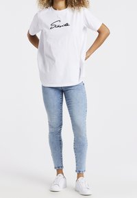 Camiseta de algodón blanca de manga corta, con un logotipo en cursiva negra. Combinada con vaqueros ajustados de color azul claro y zapatillas blancas.