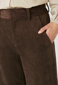 Pruunid vahuveluurist püksid vertikaalsete ribidega, millel on vöökoht ja küljetaskud, esitledes ühte kätt taskus.