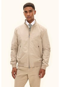 Beige Bomberjacke mit Frontreißverschluss, zwei Seitentaschen, gerippten Bündchen und glattem, leichtem Stoff. Getragen über einem weißen Hemd und einer Krawatte.