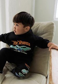Young child sitting on beige chair wearing black Hot Wheels sweatshirt and pants with car graphics, looking attentively to the left.