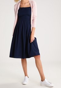 Marinblå ärmlös klänning med figurnära överdel och utsvängt kjol, parat med en ljusrosa kofta och vita sneakers. Slät tygtextur.