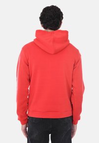 Felpa rossa in cotone con cappuccio regolabile, polsini e orlo a costine. Texture liscia. Indossata con jeans scuri, mostra un design semplice senza logo.