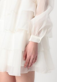 Rochie albă stratificată cu volane, material transparent și mâneci lungi bufante. Mânecile au un detaliu cu un singur nasture la manșetă.