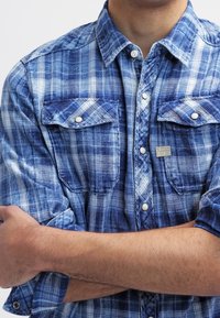 Man i blå rutiga skjorta med knappar och uppvikt ärmar, med armarna korsade över bröstet, synlig från axlarna till mitten av överkroppen.