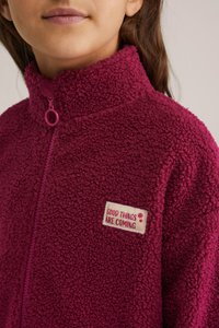 Fleece jakna v temno roza barvi z visokim ovratem, sprednjim zadrgo in aplikacijo z napisom "PRIDEJO DOBRE STVARI" izšito v kontrastnih barvah.