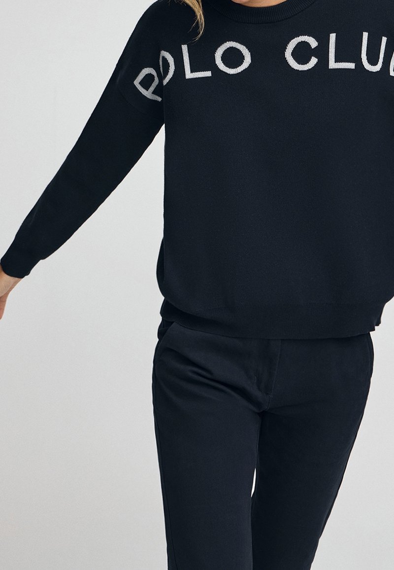 Personne portant un pull noir avec le texte « POLO CLUB » et un pantalon noir assorti sur un fond clair uni.