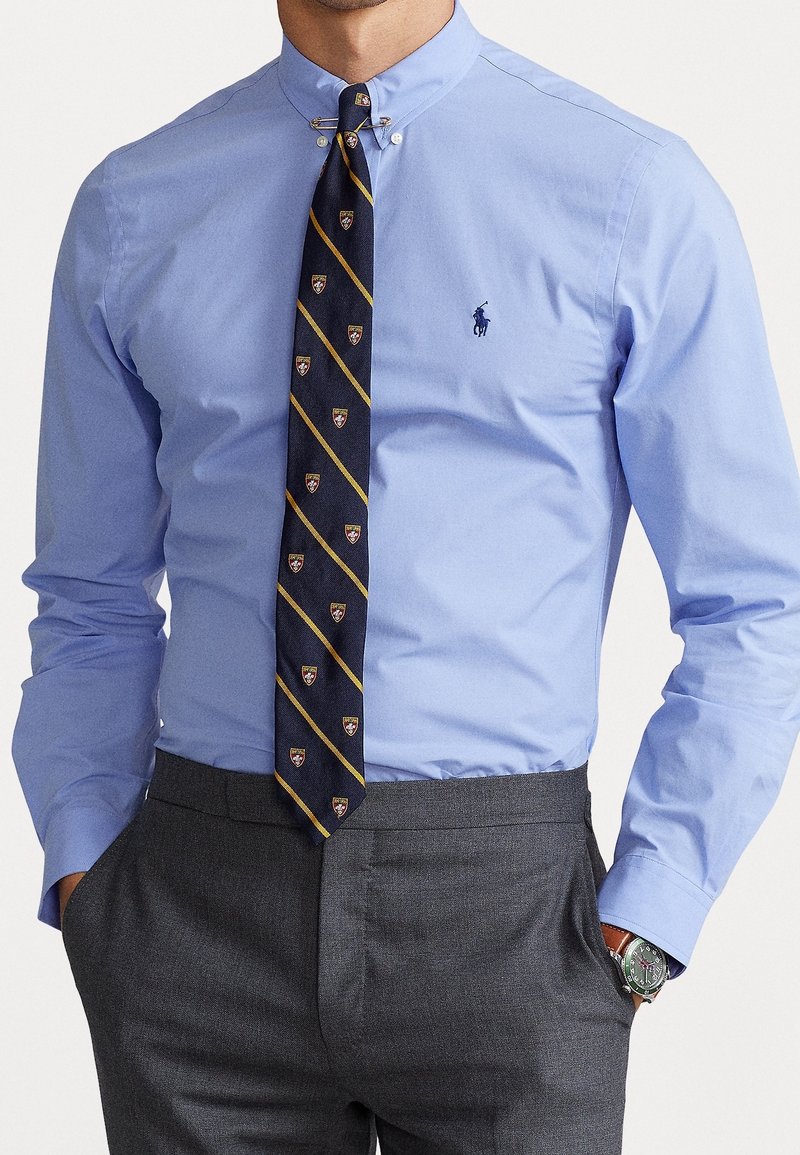 Hellblaues Button-Down-Hemd mit einer marineblauen Krawatte, die diagonale Streifen und Wappenmuster aufweist, kombiniert mit dunkelgrauen Anzughosen.
