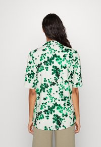 Camicia a manica corta in bianco con stampa floreale verde. Presenta un colletto e una vestibilità rilassata. Il materiale sembra morbido con una texture liscia.