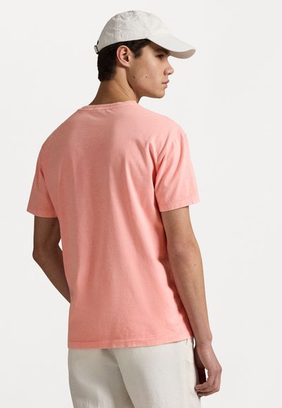 Short-sleeve, coral pink t-shirt in soft cotton, featuring a classic crew neck. Worn with a white cap and light-colored pants.