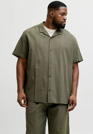 Mann som vender mot høyre, iført løs olivengrønn kortermet knapp-skjorte over hvit undertrøye og matchende bukser mot ensfarget bakgrunn.