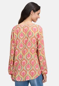 Femme aux longs cheveux noirs portant un chemisier à manches longues coloré avec des motifs floraux abstraits roses et jaunes, associé à un jean bleu.