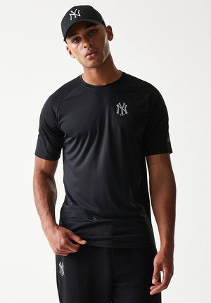 Giovane uomo che indossa un berretto nero dei New York Yankees, una maglietta sportiva nera e pantaloni neri, in piedi contro uno sfondo bianco.