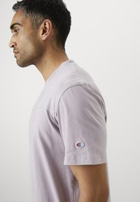 Lavenderfarbener Baumwoll-T-Shirt mit kurzen Ärmeln, rundem Halsausschnitt und einem kleinen Logo-Patch am linken Ärmel. Glatte Textur, lockere Passform.