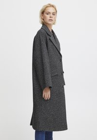 Giacca lunga in bianco e nero a motivo houndstooth con colletto a punte, chiusura a bottoni e tasche laterali, caratterizzata da una vestibilità rilassata e tessuto testurizzato.