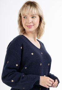 Tummansininen neulepaita, jossa on V-pääntie, kultaisia sydänkuvioita, ribbaustaskut ja pehmeä pinta.