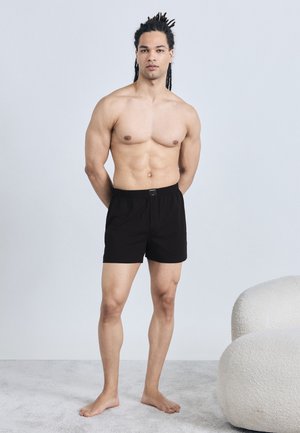 Uomo senza maglietta con dreadlocks, in piedi a piedi nudi, indossa pantaloncini neri su un tappeto chiaro accanto a una sedia bianca e arrotondata su uno sfondo semplice.