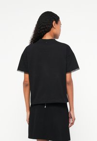 T-shirt nera a maniche corte con una vestibilità leggermente accorciata, caratterizzata da un girocollo e dettagli sovrapposti in tessuto trasparente alle maniche.