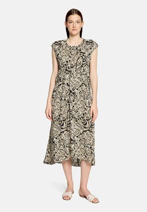 Vrouw staat, gekleed in een mouwloze midi-jurk met beige en zwart patroon en witte instapsandalen, tegen een effen lichte achtergrond.