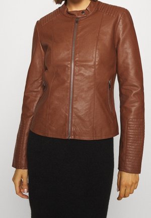 Kobieta w dopasowanej brązowej skórzanej kurtce z zamkiem błyskawicznym z przodu i kieszeniami po bokach, pikowanymi detalami na ramionach i rękawach, w połączeniu z czarną spódnicą.