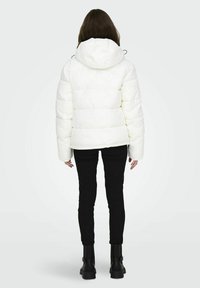 Hvid pufferjakke med hætte, indeholdende et kviltet design og brede ærmer. Kombinationen af sorte stramme bukser og kraftige sorte støvler.