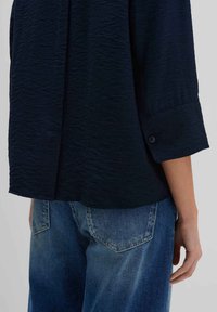Camicetta blu scuro con texture e maniche a tre quarti, dettaglio con bottoni sui polsini, abbinata a jeans di denim blu chiaro visibili da dietro.