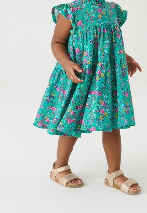 Tout-petit portant une robe turquoise à fleurs sans manches et des sandales beige, debout devant un fond clair uni.