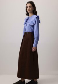 Femeie îmbrăcată cu o bluză ușor albastră, cu guler cu volane și broderie, asortată cu o fustă lungă, înaltă la talie, de culoare maro închis și pantofi negri.