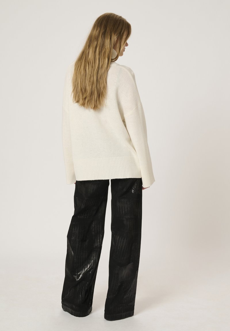 Überdimensionierter cremefarbener Strickpullover mit geripptem Saum, kombiniert mit schwarzen, strukturierten, weit geschnittenen Hosen mit glänzendem Finish und gestreiftem Muster.