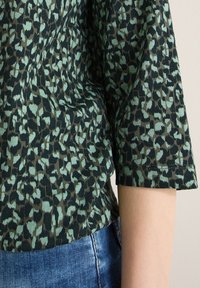 Gros plan sur un bras portant une chemise à manches courtes à imprimé léopard vert et noir, associée à un jean en denim bleu.