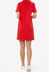 Robe rouge à manches courtes avec col, silhouette évasée en ligne A, texture lisse et ourlet à hauteur du genou. Portée avec des baskets blanches.