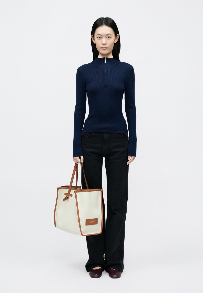 Top in maglia a coste blu navy con una zip corta, abbinato a jeans neri svasati. Tiene una borsa tote color avorio con dettagli marroni e logo.
