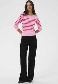 Femme debout les mains derrière le dos, portant un pull rose avec des rayures horizontales rouges et un pantalon large noir.