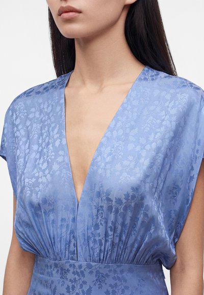 Femme portant une robe en satin bleu clair avec des motifs floraux et un décolleté en V profond, montrant le cou, les épaules et le haut du torse.