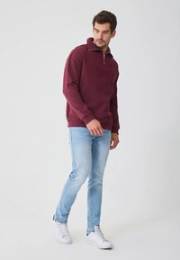 Sweatshirt borgonha com gola de fecho, em tecido macio, combinado com jeans azul-claro de corte justo e ténis brancos, posado contra um fundo neutro.