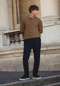 Brązowy prążkowany sweter polo z dzianiny z trzema guzikami, połączony z ciemnoniebieskimi, dopasowanymi spodniami i czarnymi tenisówkami z białymi podeszwami.