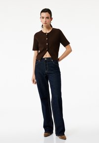 Brun kortærmet cropped cardigan med frontknapper, parret med mørkeblå flared jeans. Glatte teksturer og en afslappet pasform.