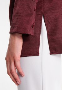 Burgundyröd långärmad skjorta med texturerad yta. Har knappdetaljer på ärmsluten. I kombination med vita byxor, med handen vilande på byxbenet.