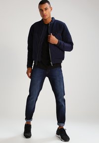 Granatowa kurtka bombowa zapinana na zamek z ściągaczami przy mankietach, w połączeniu z ciemnymi dżinsami i czarnymi trampkami. Gładki materiał, casualowy styl.