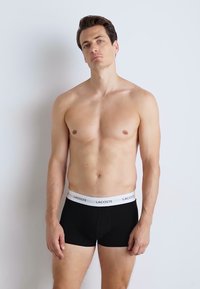 Boxers en coton noirs avec une ceinture élastique blanche arborant le logo "LACOSTE". Texture lisse, design ajusté et longueur mi-cuisse.