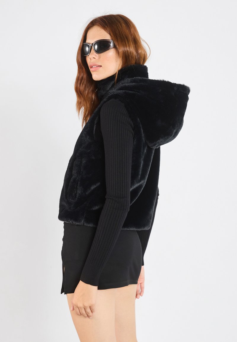 Gilet di pelliccia nera con cappuccio, caratterizzato da una trama morbida e tasche frontali. Indossato sopra un top attillato a maniche lunghe nero a coste e una mini gonna.