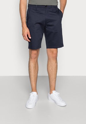 Homme portant un short bleu marine jusqu'aux genoux, une chemise grise et des baskets blanches, debout les mains dans les poches devant un fond gris.