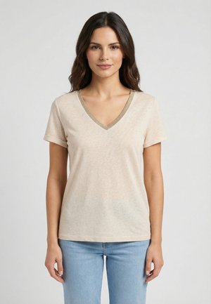 Donna con capelli castano lunghi indossa una maglietta beige a maniche corte con scollo a V e bordatura dorata, e jeans azzurri, in piedi davanti a uno sfondo semplice.