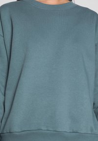 Sweat-shirt bleu sarcelle en tissu doux, avec un col rond, des manches longues et un ourlet côtelé pour un ajustement parfait. Texture lisse sans motifs.