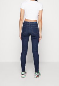 Mujer vestida con pantalones ajustados azul oscuro, una camiseta blanca corta y zapatillas gris y verde menta, de pie de espaldas.